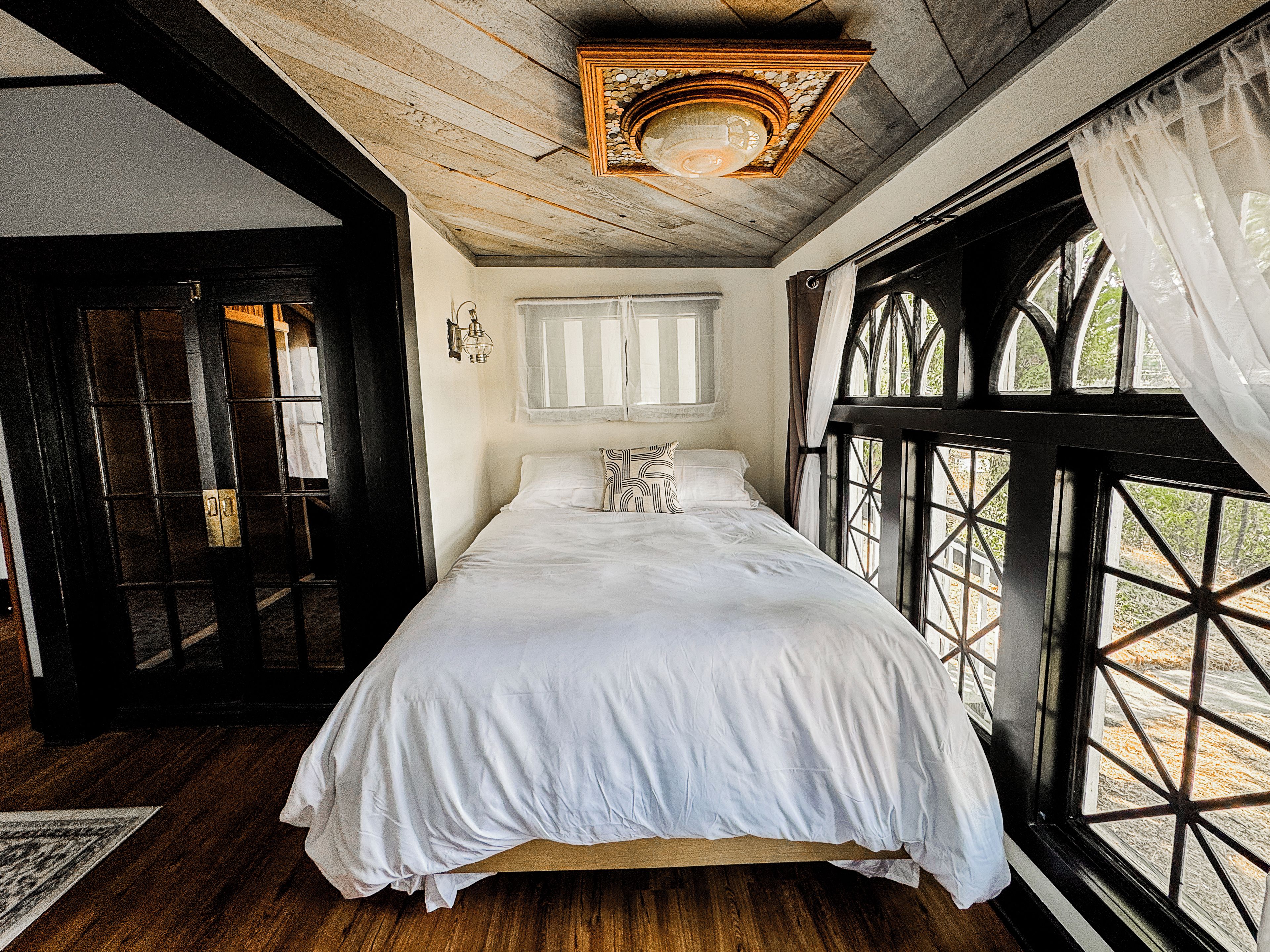 A room with a bed, a window with curtains, a wooden ceiling, and a door.