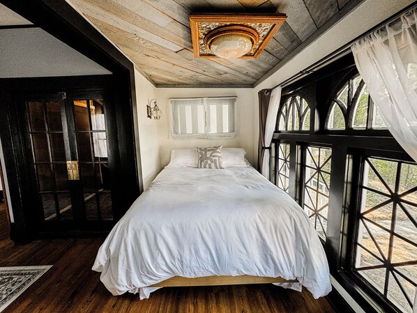Una habitación con cama, ventana con cortinas, techo de madera y una puerta.
