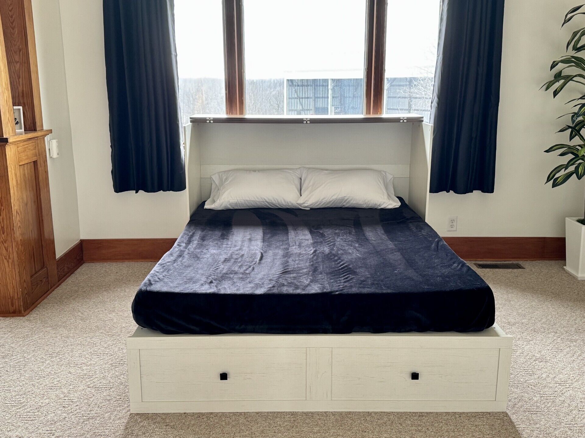 Ein Einzelbett mit Kopfteil, zwei Fenster mit Vorhängen, eine Pflanze und ein Holzschrank.