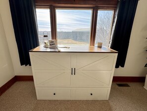 Un armario blanco de madera con dos puertas y una encimera, situado frente a una ventana con cortinas.