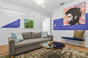 Ein modernes Wohnzimmer mit einem grauen Sofa, blauen Kissen mit Muster, einem runden Glastisch und einer blauen Bank mit Kissen.