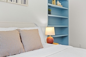Ein Bett mit karierten Kissen, eine Nachttischlampe und ein blaues Regalsystem an einer weißen Wand.