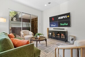 Ein modernes Wohnzimmer mit Sofa, Sessel und Couchtisch. Ein Kamin ist vorhanden, darüber ist ein Fernseher angebracht, der die Logos von Netflix, ESPN+, Disney+ und Hulu zeigt.