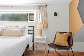Ein Schlafzimmer mit Bett, Nachttisch, Lampe, Stuhl und Blick auf Bäume durch das Fenster.