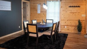 En matplats med ett runt bord, sex stolar, en svart matta, träväggar, ett fönster med persienner och en liten krukväxt.