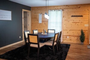 A dining area with a round table, six chairs, a black rug, wooden walls, a window with blinds, and a small potted plant. - 3BR/2BA • 4-6 Min to Huzzah Valley & Bass River Resort (Steelville)