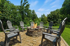 Ein Steinkamin mit vier Adirondack-Stühlen, die darum angeordnet sind, auf einer gepflasterten Terrasse mit einer Rasenfläche und Bäumen im Hintergrund.