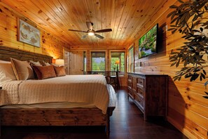 A wooden cabin interior with a bed, a ceiling fan, a television, and a view of the outdoors through a window. - Peaceful Wooded Retreat - Arcade, Hot Tub + Fire Pit (Sevierville)