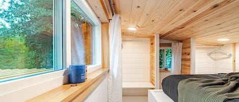 A cabin interior with a bed, a window with a view of trees, and a ceiling with wooden beams.