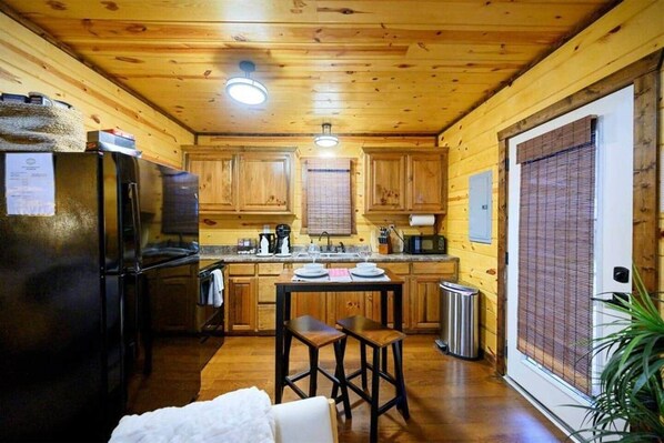 A compact kitchen with wooden cabinets, a refrigerator, a sink, and a small dining area with stools. - Beaver's Little Lodge I Couples Cabin I Romantic (Hochatown)