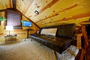 Ein gemütliches Schlafzimmer im Dachgeschoss mit einem Ledersofa, einem kleinen Tisch mit Lampe und einem Fernseher an einer Holzwand.