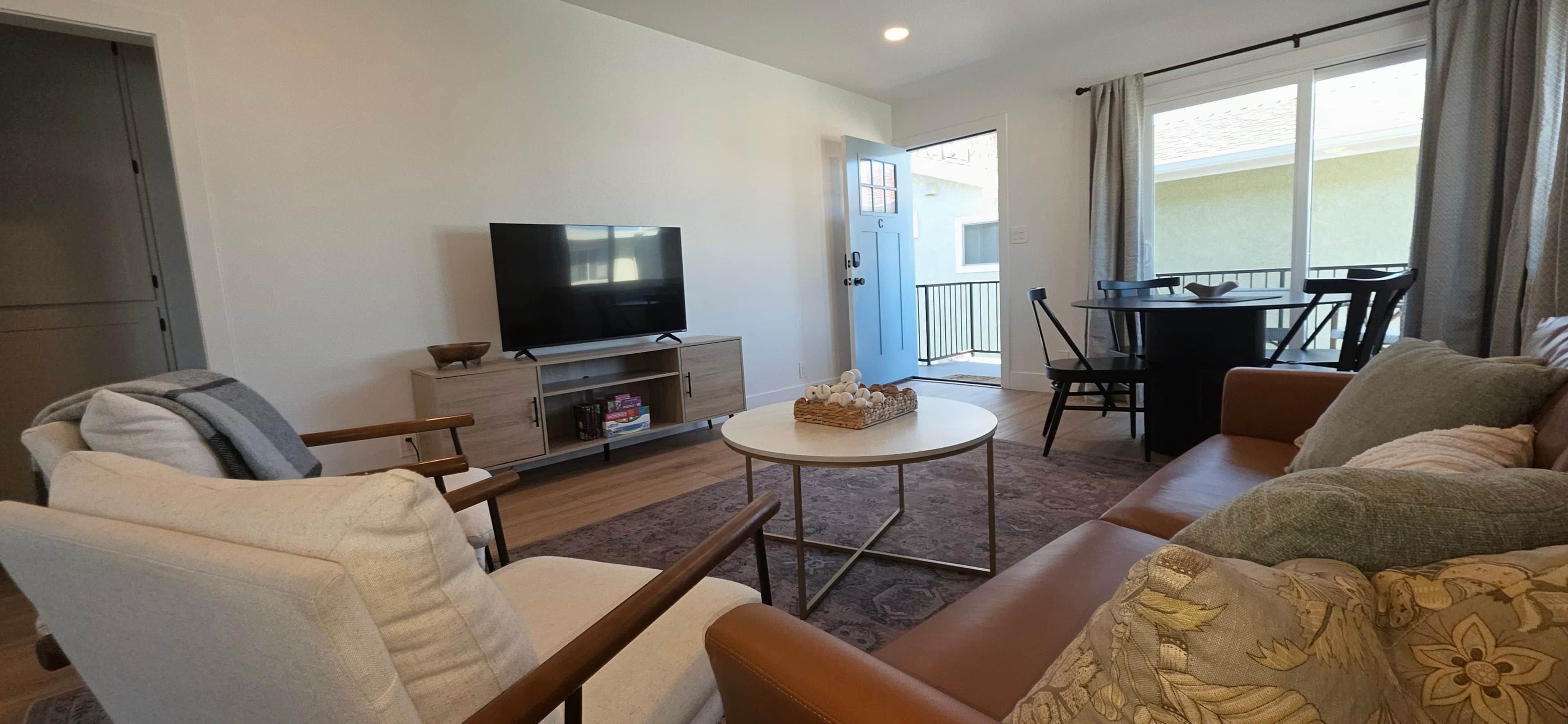 A living room with a flat-screen TV, a sofa, a chair, a small table, and a balcony with a dining table and chairs.