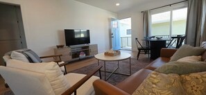 A living room with a flat-screen TV, a sofa, a chair, a small table, and a balcony with a dining table and chairs. - Newly Renovated Coastal Condo with Peekaboo Ocean View (San Clemente)