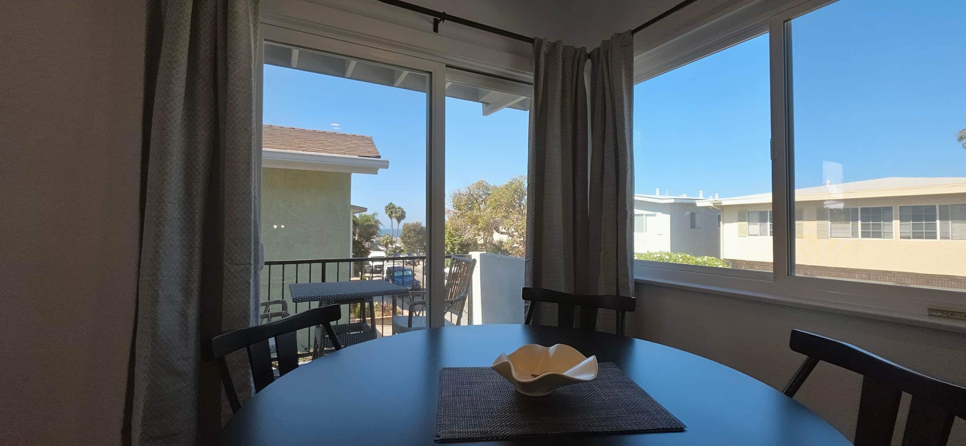 A room with a table and chairs, a window with curtains, and a view of buildings and trees outside.