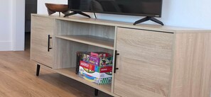 A wooden TV stand with a flat-screen TV on top, a shelf below, and a box of Monopoly games on the lower shelf. - Newly Renovated Coastal Condo with Peekaboo Ocean View (San Clemente)