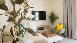 Ein modernes Wohnzimmer mit Fernseher, Glastisch, Sofa, Sessel mit gelber Kissen und Topfpflanze.