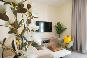 Ein modernes Wohnzimmer mit Fernseher, Glastisch, Sofa, Sessel mit gelber Kissen und Topfpflanze.