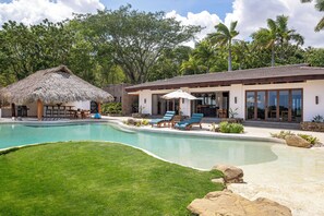 Ein Poolbereich mit Liegestühlen, einer Struktur mit Reetdach und einem modernen Gebäude mit großen Fenstern.
