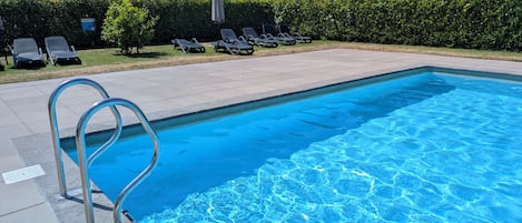 A clear blue swimming pool with a metal ladder, surrounded by a paved deck and lounge chairs.