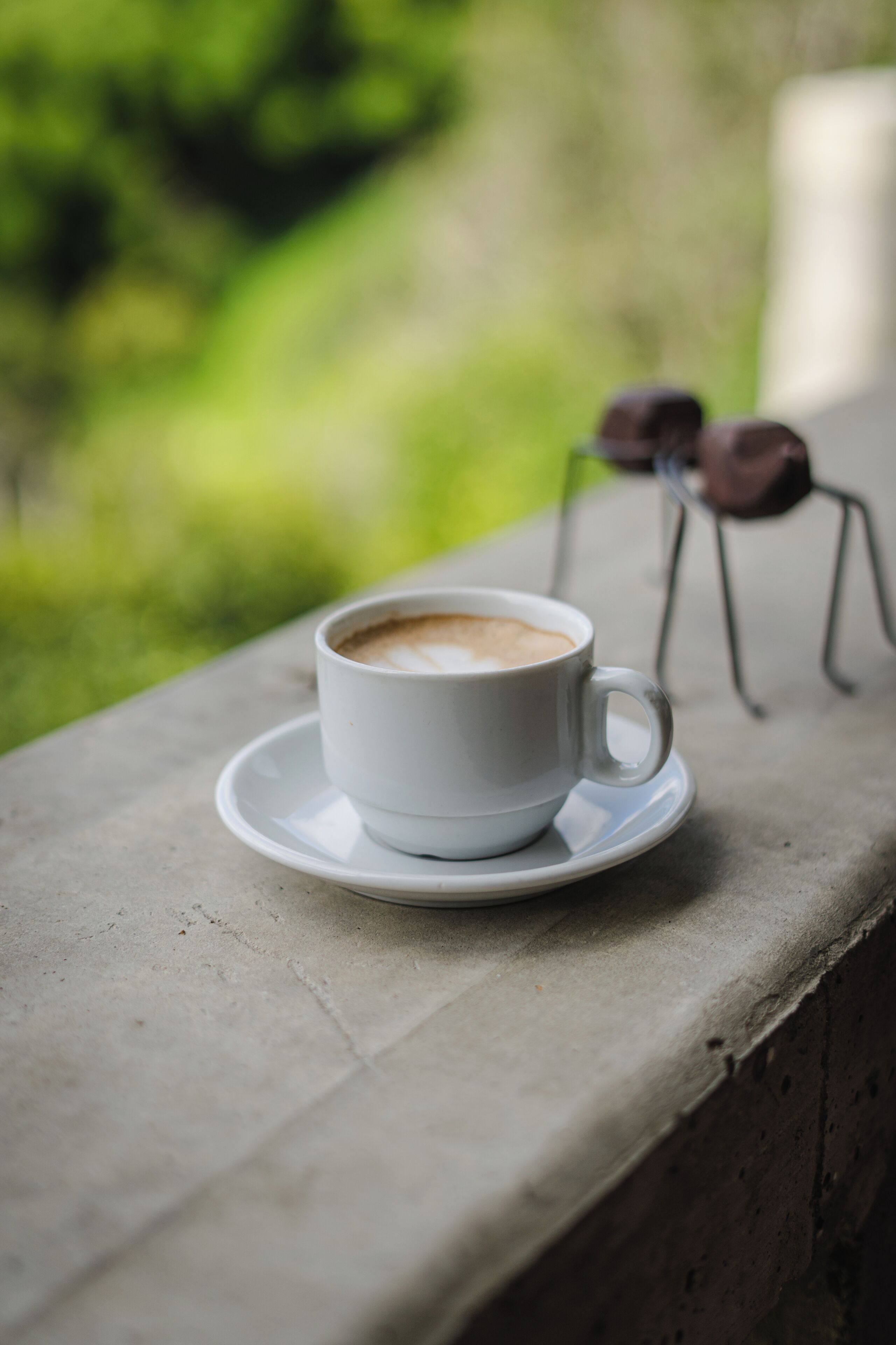 A white cup of coffee on a saucer placed on a concrete ledge with a blurred green background.