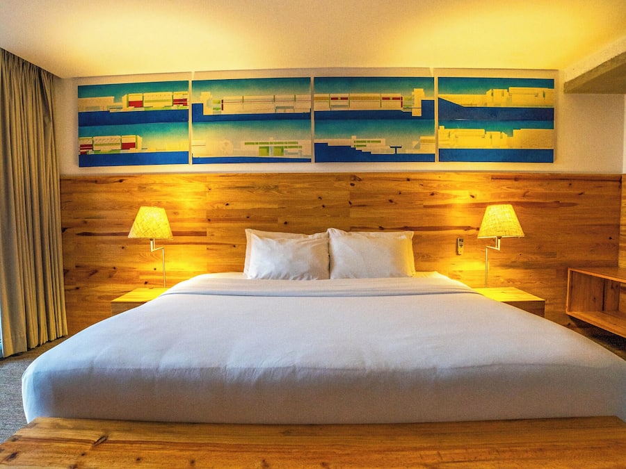 Une chambre d’hôtel avec un grand lit, une tête de lit en bois, deux lampes de chevet et un mur orné d’œuvres d’art abstraites.