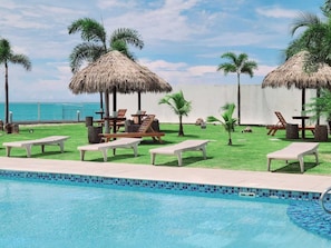 Zona de piscina con sillones de descanso, sombrillas de paja y vista al océano.