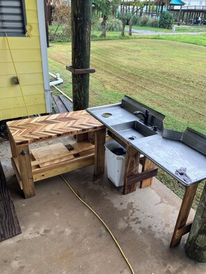 Een houten werkbank met een spoelbak en een prullenbak, op een betonnen oppervlak met een gazon op de achtergrond.