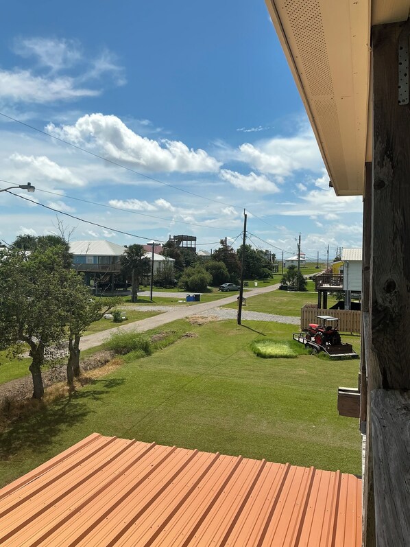 A view from a balcony with a wooden floor, overlooking a grassy area with a tractor and a car, and buildings in the distance.