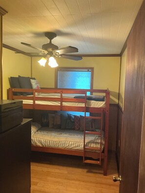 Een slaapkamer met stapelbed, plafondventilator, raam met zonwering en een deur.