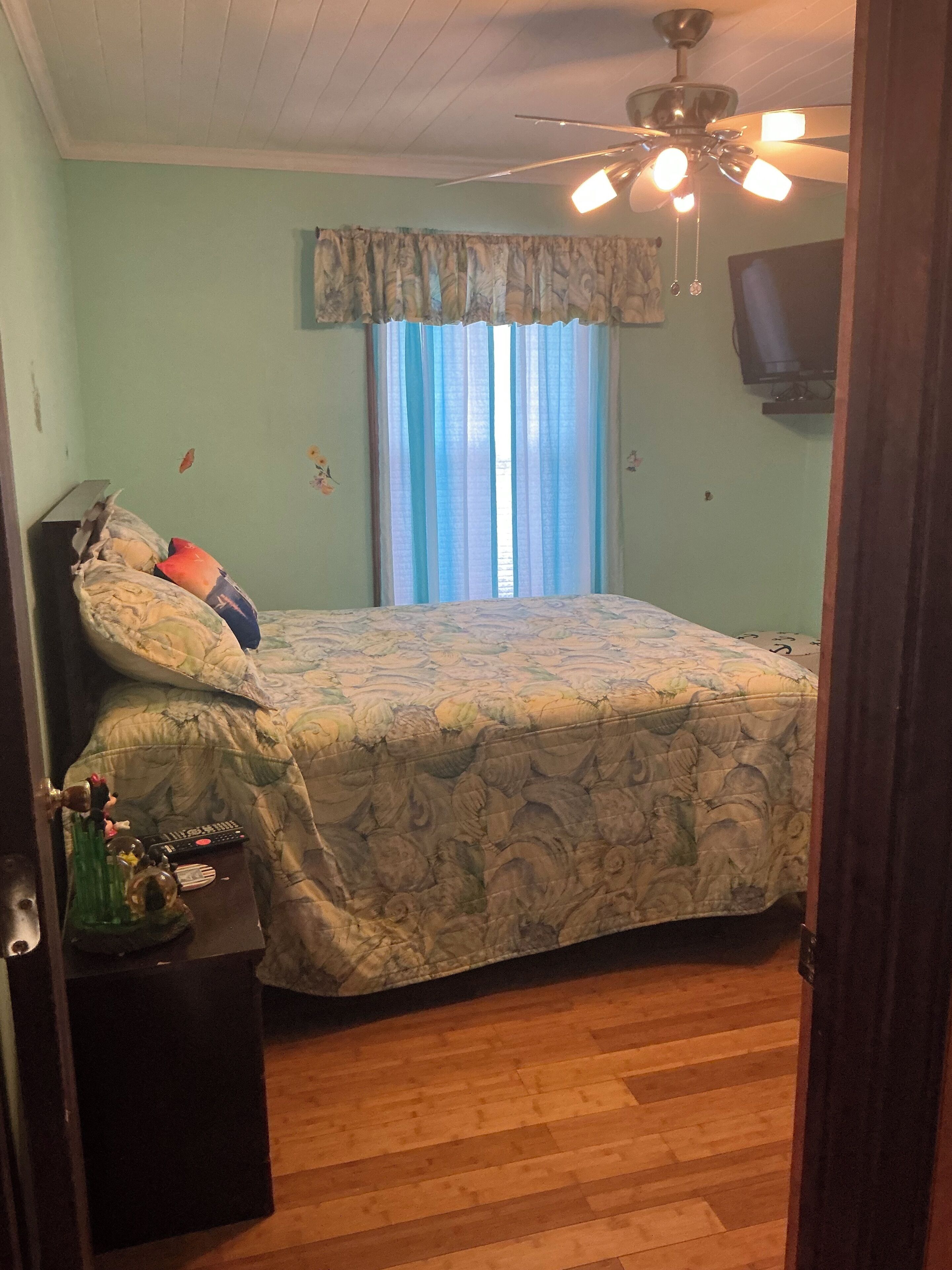 Une chambre à coucher avec un lit, un ventilateur de plafond, une fenêtre avec des rideaux et un téléviseur fixé au mur.