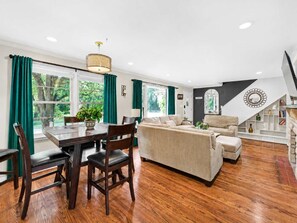 A modern living room with a dining area, wooden flooring, and green curtains. - Casa Newburg | Hot Tub • Game Garage • Fenced Yard (Louisville)