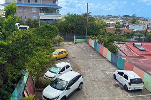 A parking area with multiple cars, surrounded by greenery and buildings in the background. - Le petit havre de paix - Climatisation (Schœlcher)