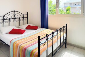 A bed with a metal frame, white bedding, and a colorful striped blanket. - Le petit havre de paix - Climatisation (Schœlcher)