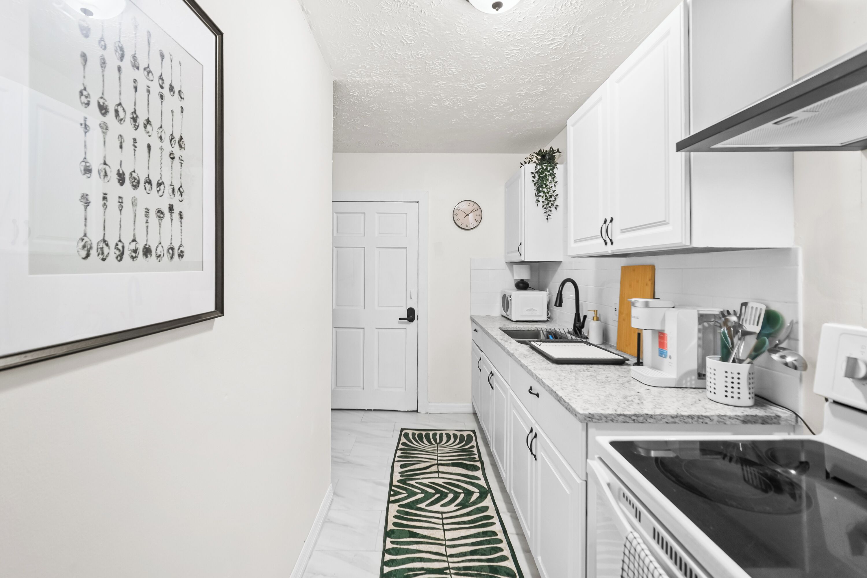 Une cuisine avec des meubles blancs, un four à micro-ondes, une bouilloire et une cuisinière. Un tableau encadré représentant des cuillères est accroché au mur.