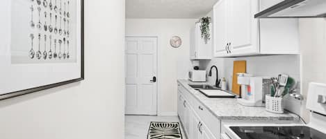 Una cocina con armarios blancos, microondas, hervidor y estufa. Hay un cuadro con cucharas en la pared.