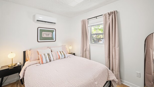 Een slaapkamer met een bed, nachtkastjes, een airconditioningapparaat, een raam met gordijnen, en een bord met een lijst op de muur.
