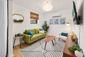 Un salon avec un canapé vert, un canapé blanc, une table basse en bois, un tapis à rayures et un luminaire au plafond.