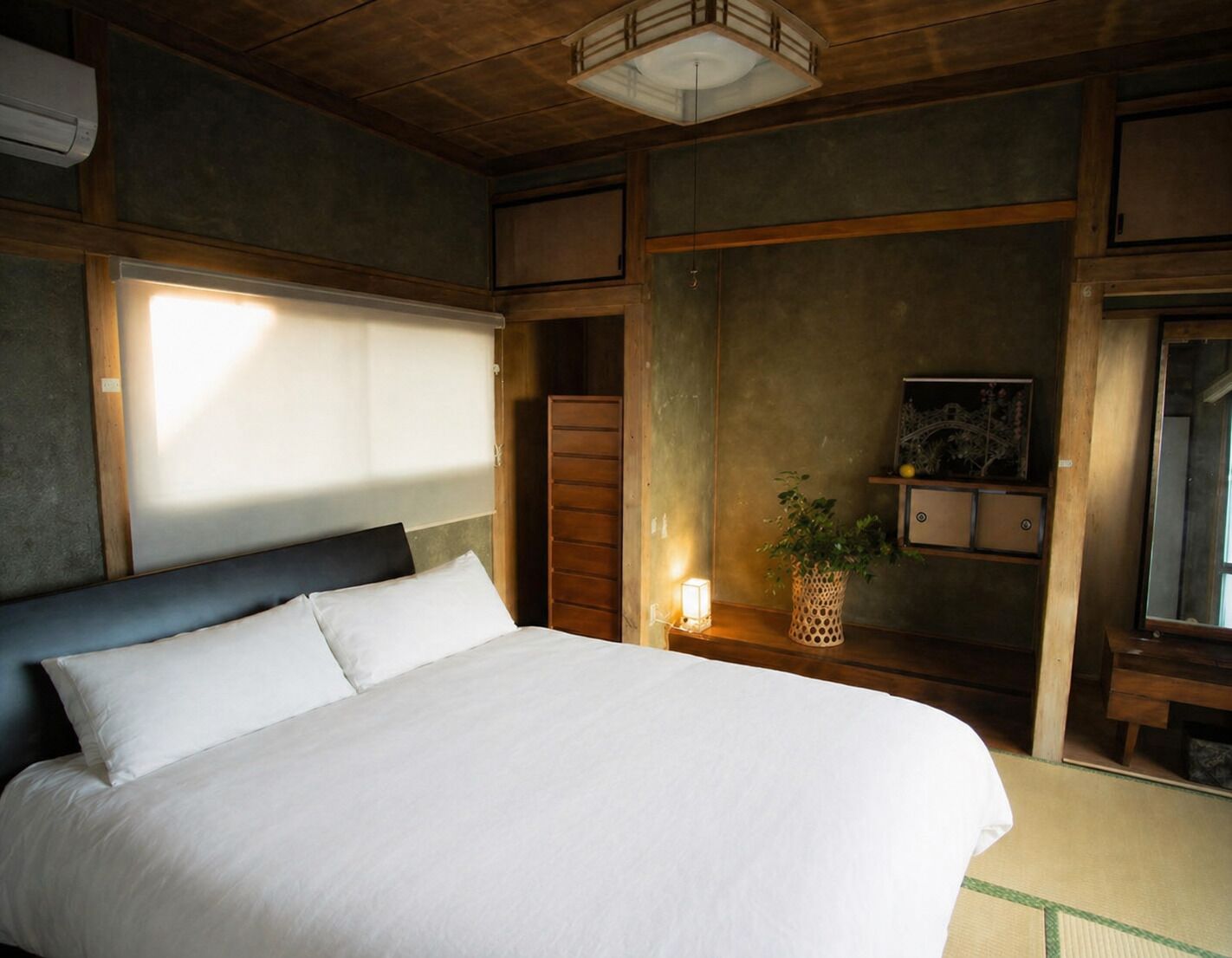 A bedroom with a bed, a bedside table with a lamp, a wall-mounted shelf with decorative items, and a window with blinds.