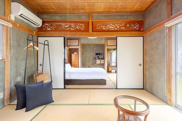 A traditional Japanese-style room with tatami flooring, a bed with white bedding, a hanging wardrobe, and a small table with a chair.