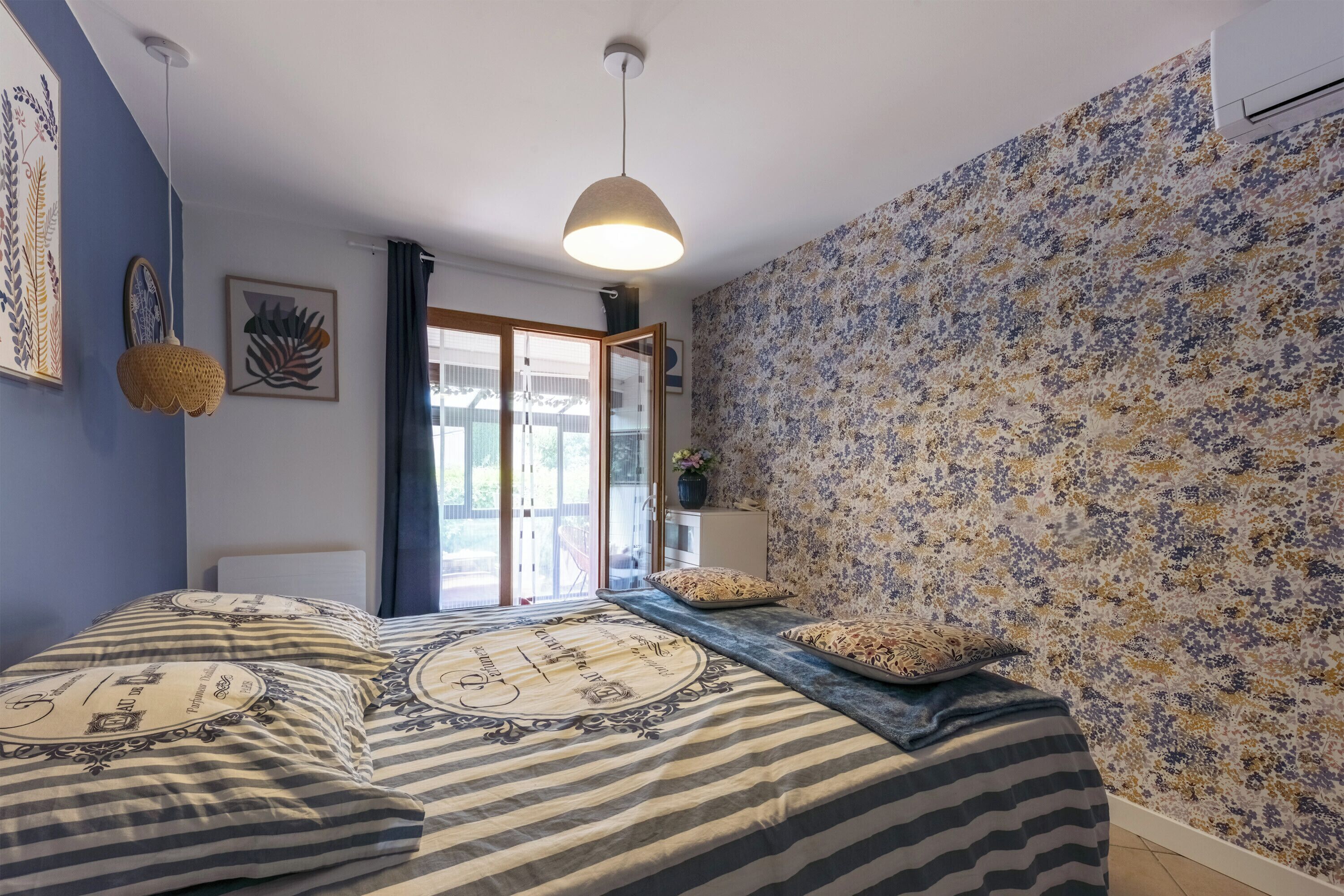 A bedroom with a bed, a hanging light fixture, a window with curtains, and a floral-patterned wall.