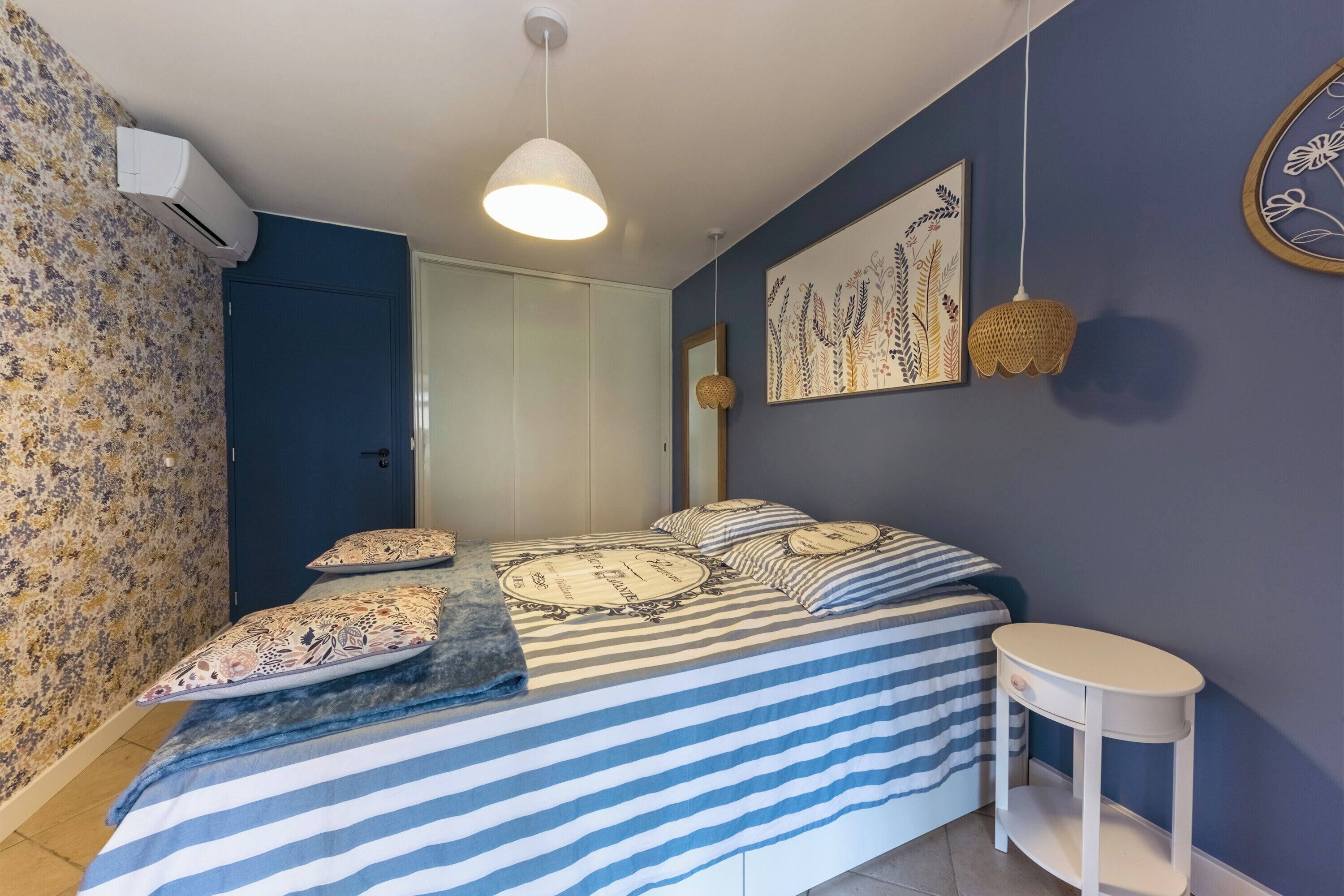 A bedroom with a bed, a bedside table, a hanging light fixture, a wall-mounted artwork, and an air conditioning unit.