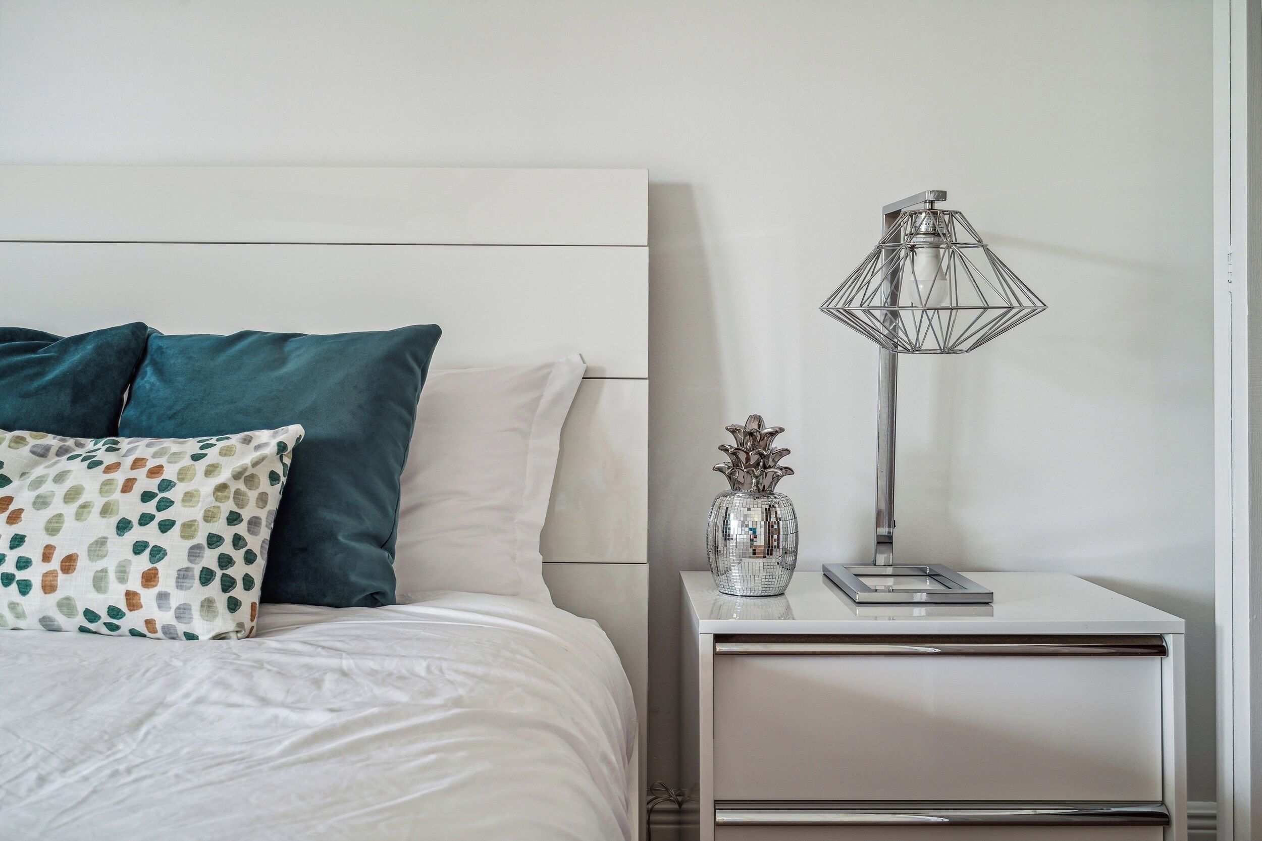 A neatly made bed with white and teal pillows, a small bedside table with a metallic lamp and a decorative vase, and a plain wall.