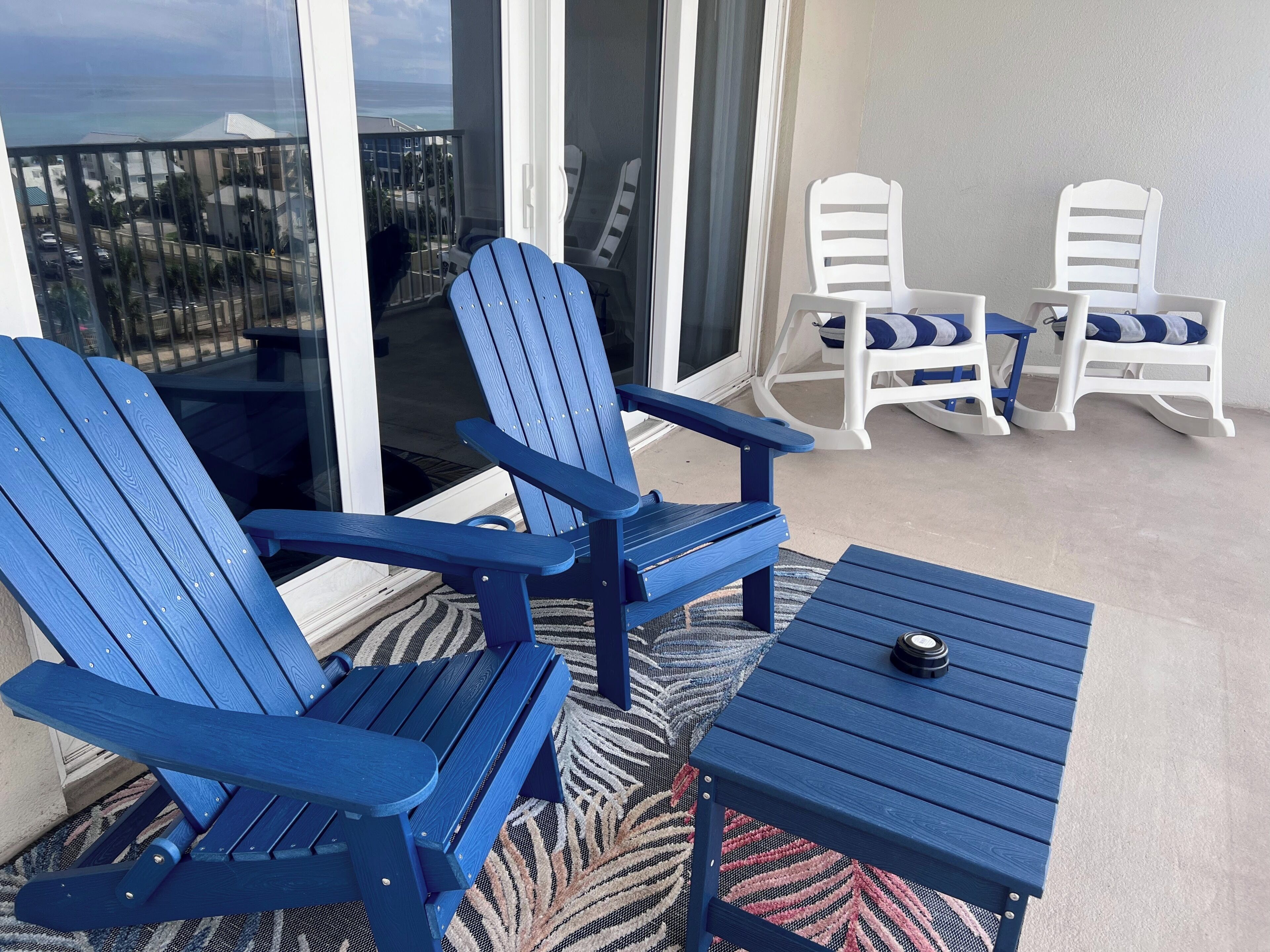 A balcony with two blue Adirondack chairs, a small table, and white rocking chairs.