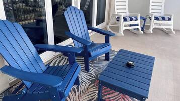 A balcony with two blue Adirondack chairs, a small table, and white rocking chairs.