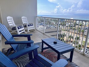Un balcon avec des chaises bleues et une table, donnant sur une rue et l’océan.