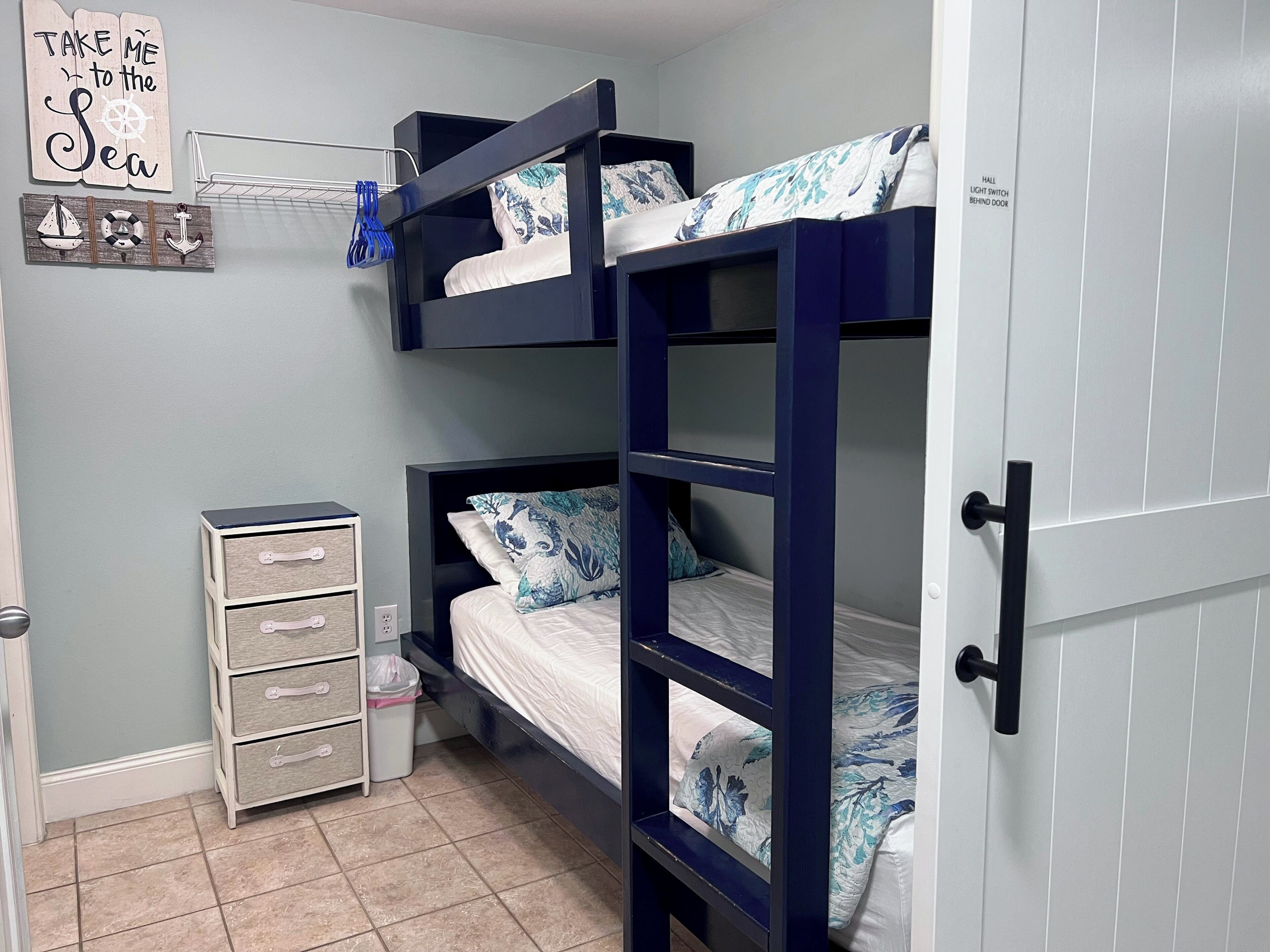 A bunk bed room with a small dresser, a wall-mounted shelf with nautical decorations, and a door with a light switch label.