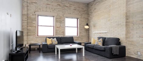 A modern living room with a brick wall, two windows, a sofa set, a coffee table, a TV, and a floor lamp.