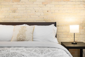Una cama con una colcha estampada y dos almohadas, una con diseño de piña, frente a una pared de ladrillo. Se observa una mesita de noche con una lámpara.