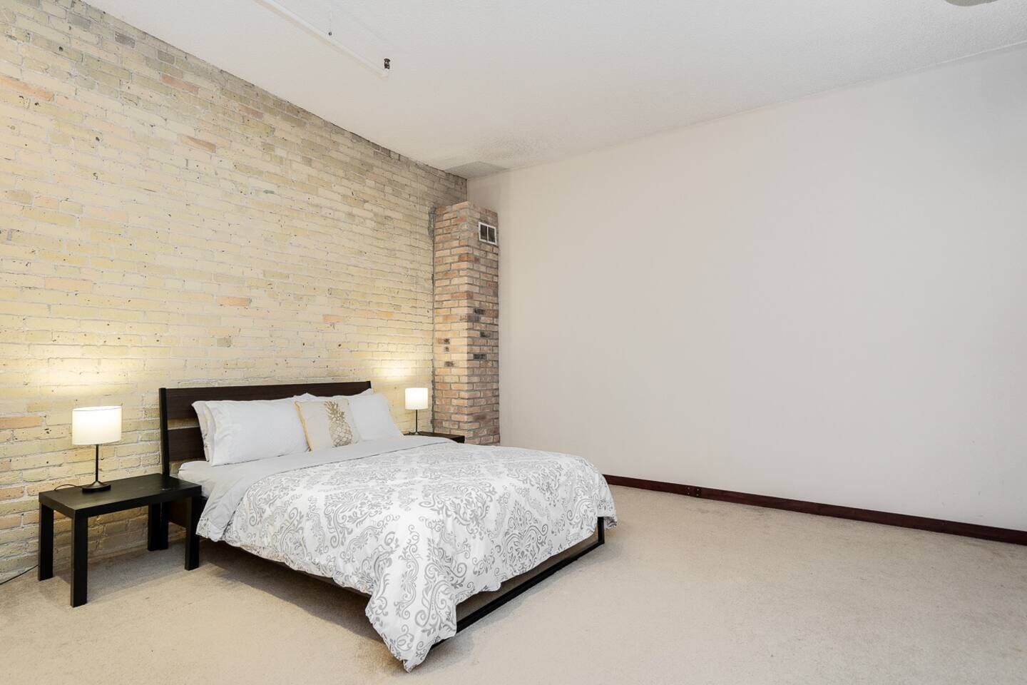 Un dormitorio con pared de ladrillo, una cama con colcha estampada, dos lámparas de noche y un pequeño pilar de ladrillo.
