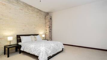 A bedroom with a brick wall, a bed with a patterned bedspread, two bedside lamps, and a small brick column.